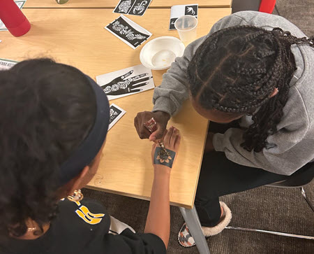 A student carefully applies henna paste to another a flower stencil on another student's hand