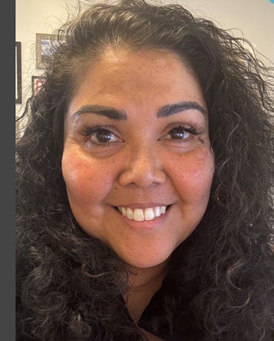 Close, self portrait of Priya Pandya-Orozco, a brown-skinned woman with curly, dark brunette hair. In the backgroun, one can just make out an office space.