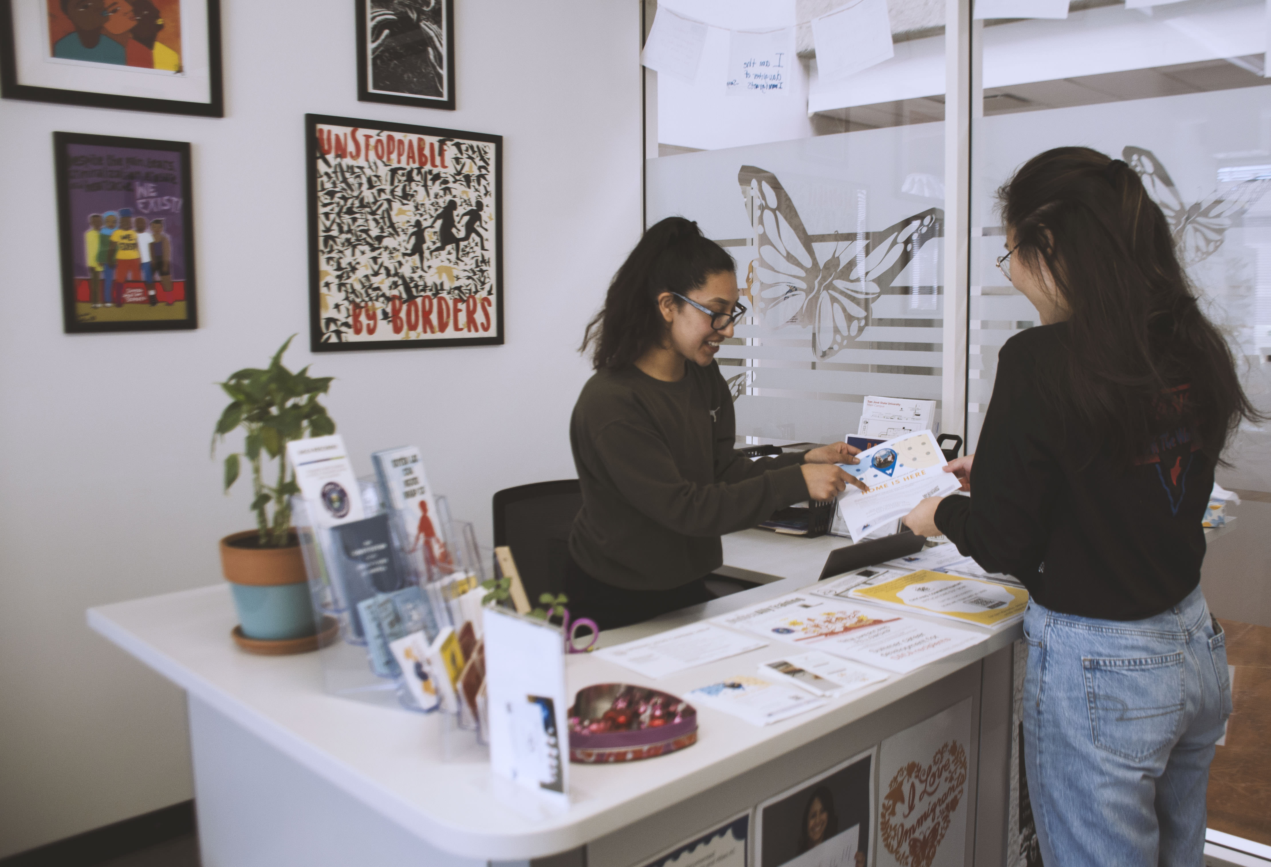 Front desk area of the UndocuSpartan Student Resource Center at San Jose State University, featuring two young students at the front desk with informational materials, a welcoming atmosphere, and a backdrop with decorations related to the center's mission.